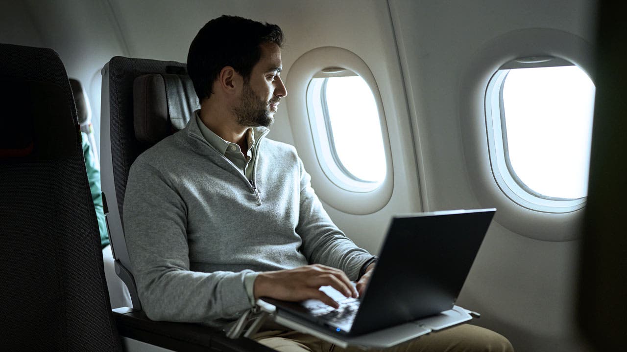 A man sitting in an airplane seat, next to two windows. He is wearing a light gray coat and has a laptop open on the table in front of him. The passenger looks pensively out of the window while using the computer, in an environment illuminated by the natural light that enters the plane.