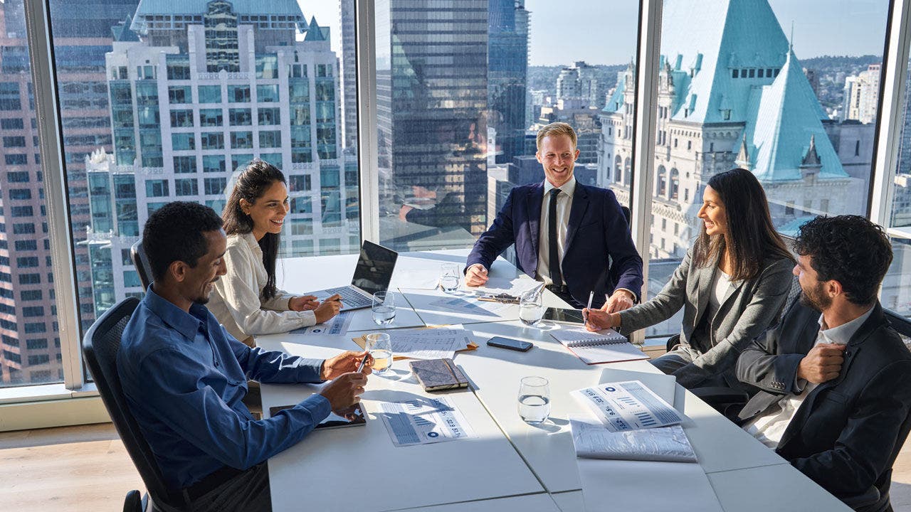 Group of five professionals smiling during a meeting in a modern conference room with panoramic windows and a view of skyscrapers. There are documents, glasses of water, laptops and cell phones on the table. Natural light illuminates the space, suggesting a collaborative and positive environment.