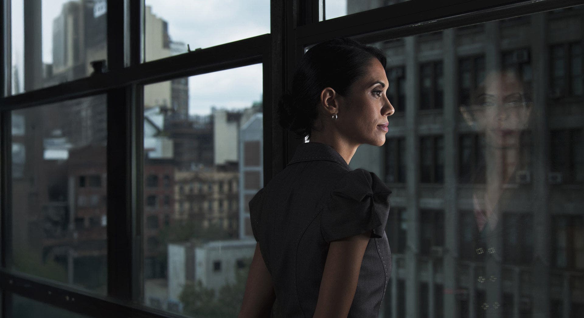 A woman seen from behind, standing by a large window, looks outside. She is wearing a dark short-sleeved blouse or dress. Outside, there is an urban scene with tall buildings and windows, suggesting that the photograph was taken in a city.