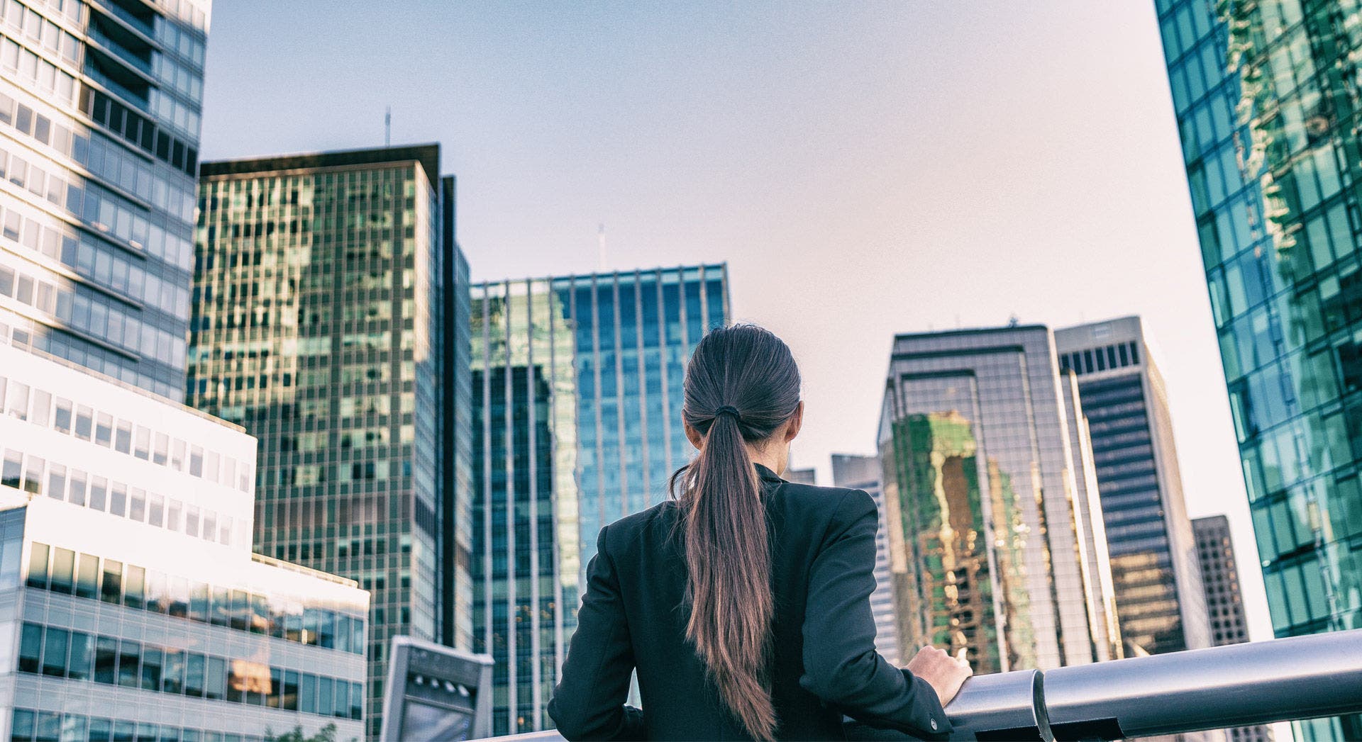 Donna con i capelli legati, vista di spalle, che guarda alti edifici per uffici in un quartiere aziendale moderno.