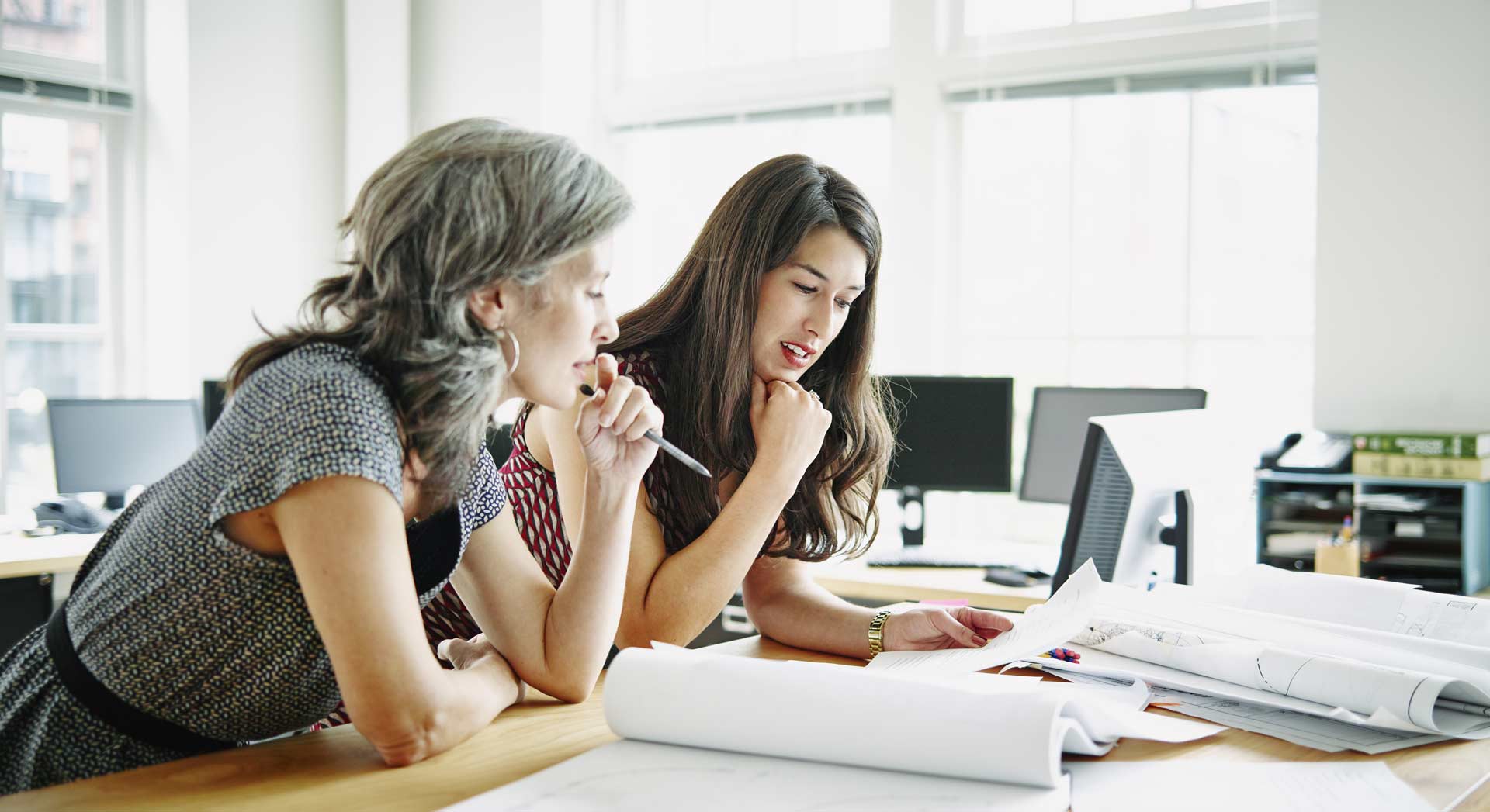 Zwei Frauen sitzen nebeneinander an einem Schreibtisch in einem modernen, gut beleuchteten Büro mit großen Fenstern im Hintergrund. Beide sind konzentriert damit beschäftigt, Dokumente und technische Zeichnungen bzw. Pläne zu analysieren, die auf dem Tisch ausgebreitet sind. Eine der Frauen hält einen Stift und im Hintergrund sind Computermonitore und Büromöbel zu sehen.