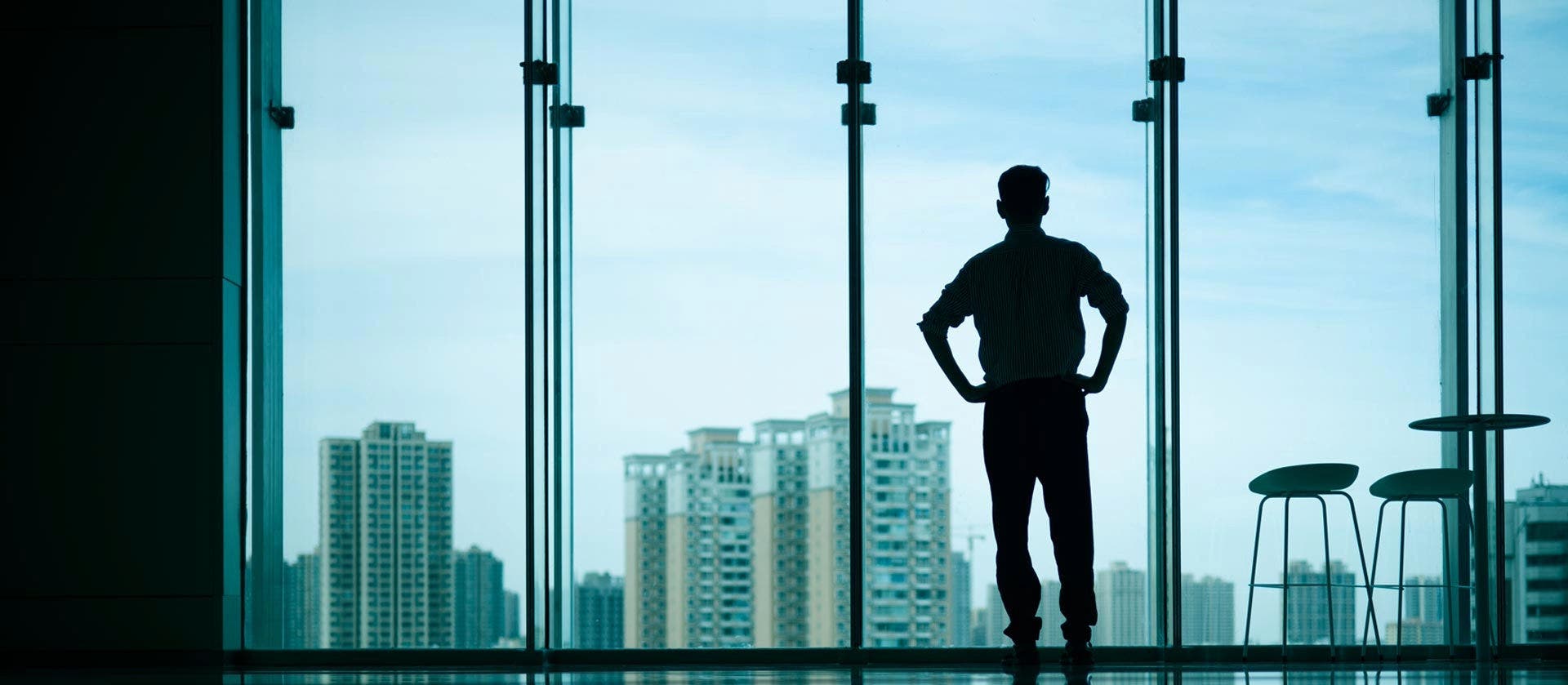Silhouette of a man standing, with his back to the camera, next to a large panoramic glass window. With his hands on the waist, he looks outside, where several tall buildings can be seen in the background.  To the right of the man are two tall chairs and a table.