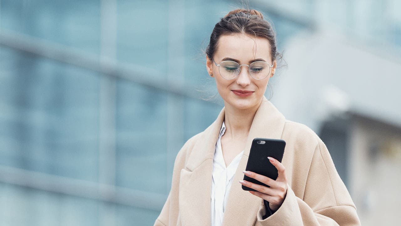 Image of a brunette wearing glasses, a beige coat, and a white shirt. The woman is looking at the cell phone she holds in her left hand. In the background, large windows suggest the setting is a corporate building.