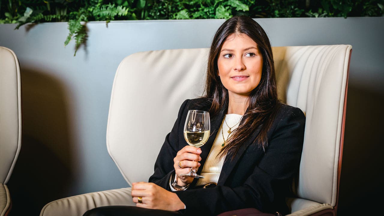 A woman sitting in an armchair, in a modern environment. She wears a black blazer over a light-colored top and holds a glass of white wine in her right hand. The background features a gray wall with decorative plants on top.